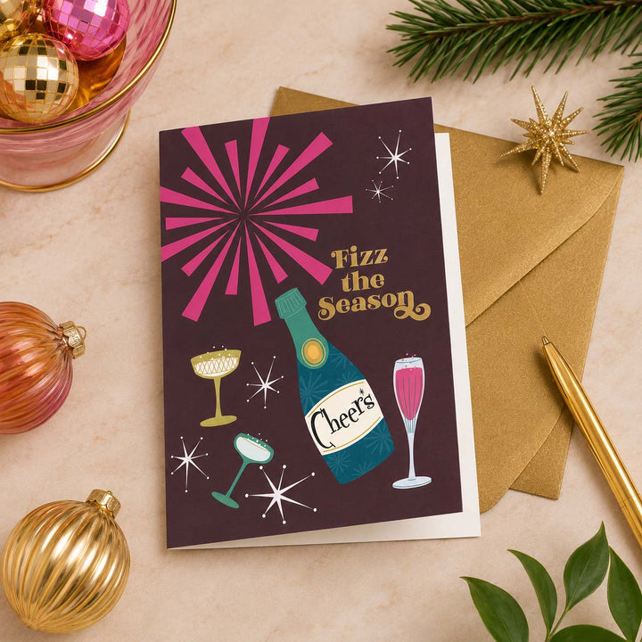 Fizz the Season holiday greeting card displayed partially open with an antique gold Euro flap envelope, surrounded by vintage Christmas ornaments and festive cocktail party décor.