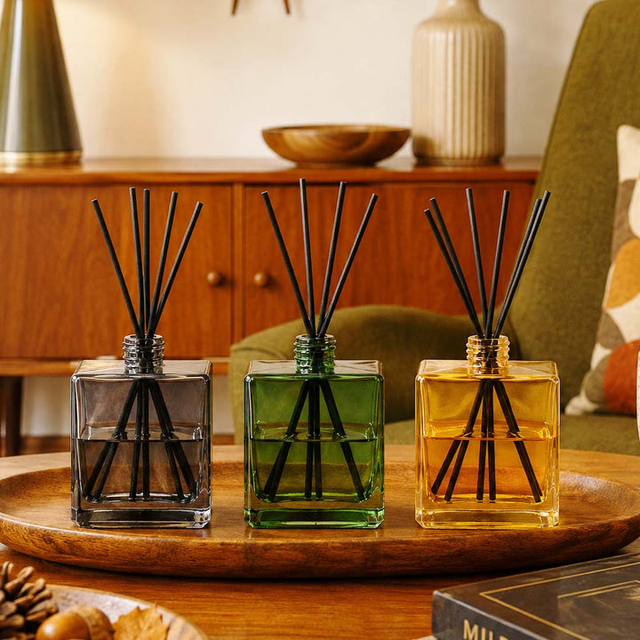 Three glass reed diffusers with black reeds, one smoky gray, one translucent green, and one golden yellow, arranged on a wooden tray in a warm, styled room setting.
