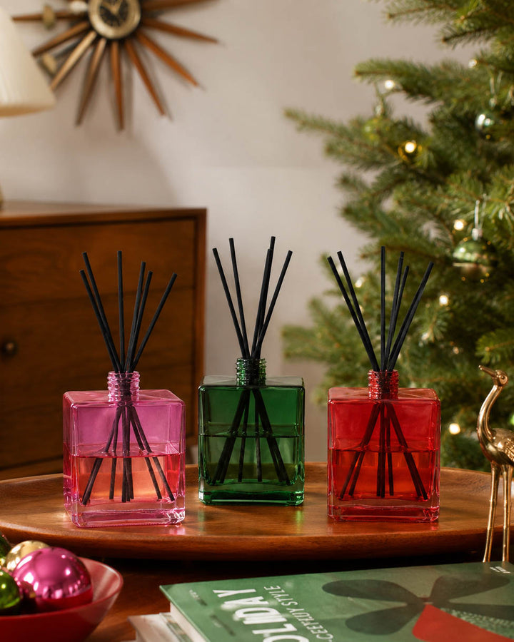 Three reed diffusers in pink, green, and red glass bottles with black reeds, arranged on a wooden tray in a cozy indoor setting with a Christmas tree in the background.