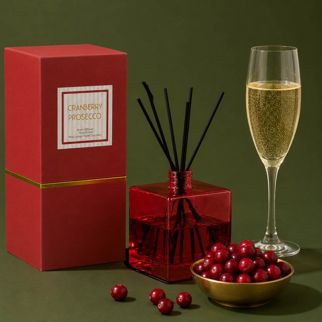 A cranberry prosecco reed diffuser set on a dark olive green background, featuring a red gift box with gold trim, a red glass diffuser bottle with black reeds, a gold bowl of fresh cranberries, and a filled champagne flute.