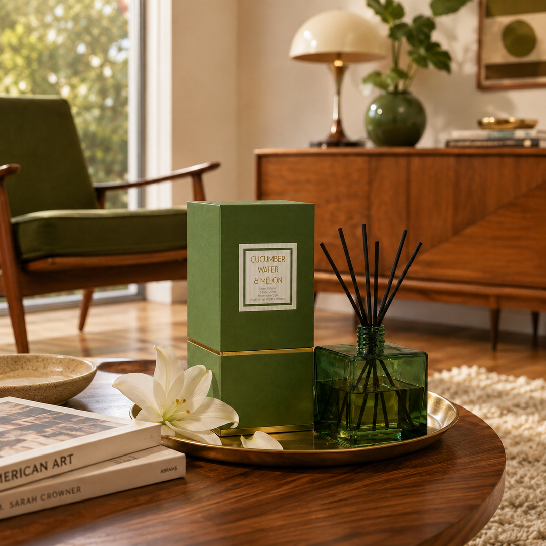 A green reed diffuser box and matching glass diffuser bottle with black reeds sit on a gold tray on a wooden table in a styled living room. The diffuser box has a white label reading &