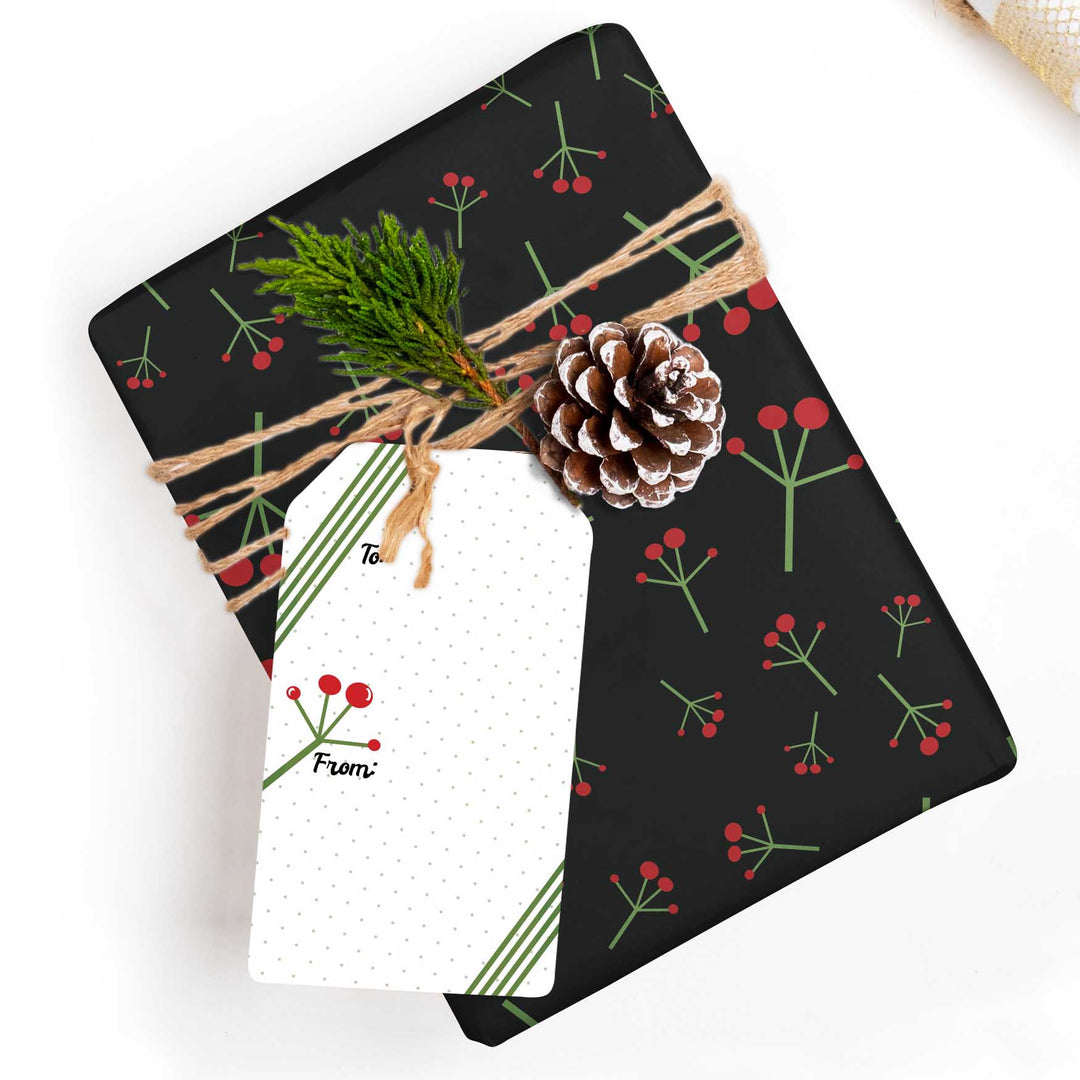 A wrapped gift box with a black holiday-themed wrapping paper featuring a red berry and green stem pattern, tied with twine and topped with a pinecone, evergreen sprig, and a white gift tag.