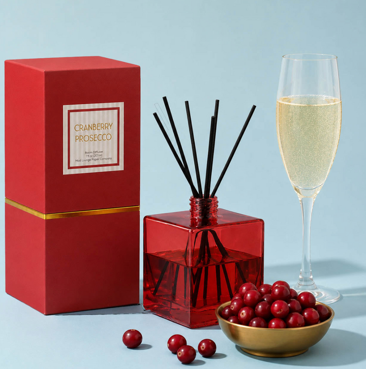 A cranberry prosecco reed diffuser set on a light blue background, featuring a red glass diffuser bottle with black reeds, a red gift box labeled &