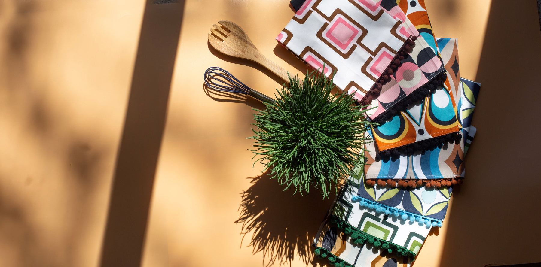 A styled flat-lay of colorful patterned napkins with black tassel edges, a small green potted plant, and wooden kitchen utensils on a warm beige surface with strong diagonal shadows.